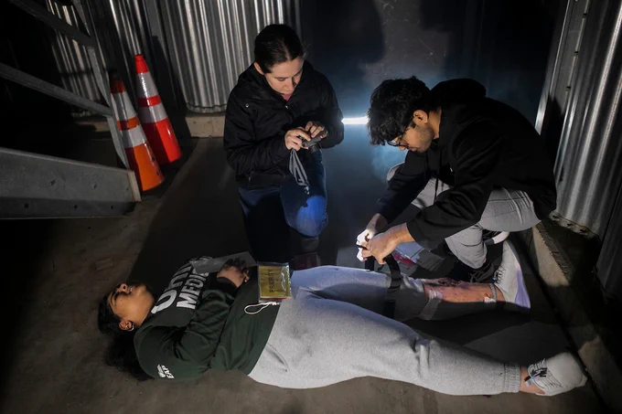 Two students provide medical aid to a simulated victim in a dark, realistic disaster training environment.