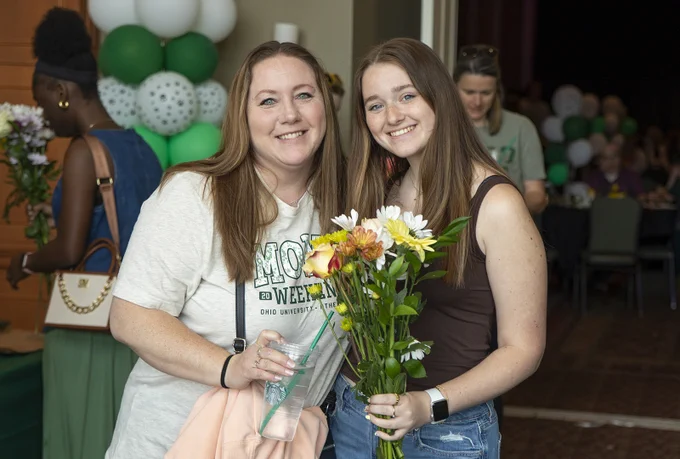Two people are smiling and standing indoors, holding flowers. They are at an event decorated with green and white balloons. One person is wearing a shirt with the text "Mom's Weekend Ohio University." There are others in the background.