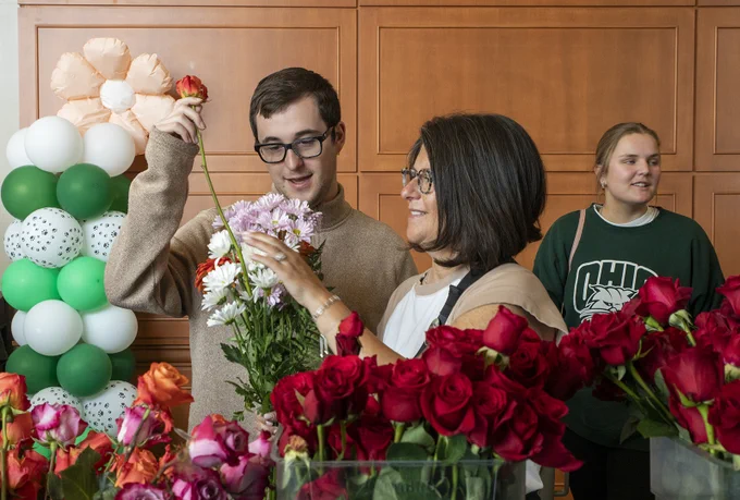 A person arranges a variety of flowers with assistance from another individual. They are surrounded by red roses and other colorful blooms. In the background, a person in an Ohio University shirt stands near a balloon display with white and green balloons.