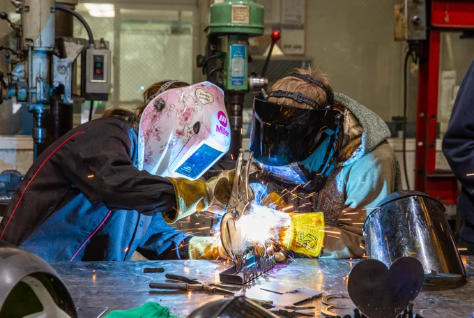 Two people in protective gear are welding at a workbench in a workshop. One has a floral welding helmet. Sparks fly from the welding area, and various tools and metal pieces are scattered on the table. Machinery is visible in the background. This is happening during a demo day for high school students to check out RCC.