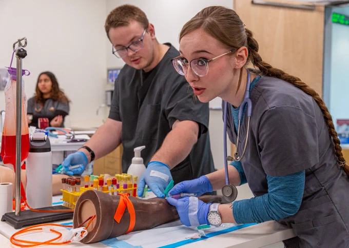 Two students in medical scrubs are practicing on a prosthetic limb in a clinical setting, using medical equipment and supplies. A laboratory test tube rack is nearby, holding various vials and fake blood. Another person is visible in the background. This is happening during a demo day for high school students to check out RCC.