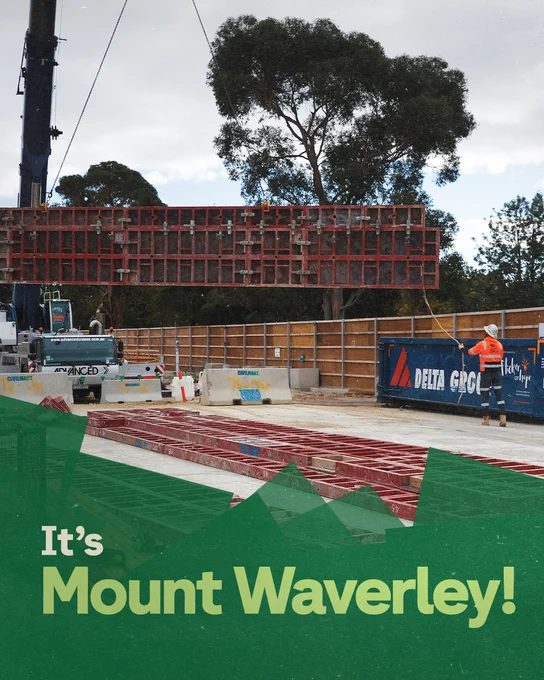 A construction site at Mount Waverley is shown. A crane is lifting a large section of wall framing. A person is guiding the operation. Construction barriers display the Delta Group logo. In the foreground, text reads, "It's Mount Waverley!" Trees and a cloudy sky are visible in the background.