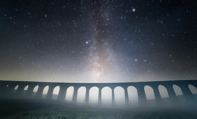 Starry night sky with the Milky Way visible above a long, arched stone aqueduct shrouded in mist.
