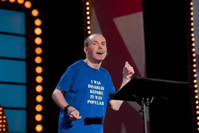 Lee Ridley (Lost Voice Guy) performing stand-up on stage, wearing a blue T-shirt that reads “I was disabled before it was popular.” He stands beside a communication device on a stand, mid-performance, with stage lights behind him.