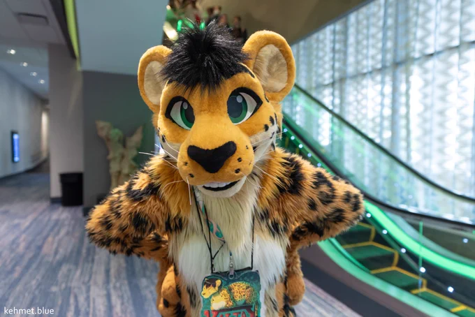 A photograph taken in front of the escalators at the Hyatt Regency. A big cat fullsuiter is leaning towards the camera with their hands on their hips. A watermark in the corner says kehmet.blue.