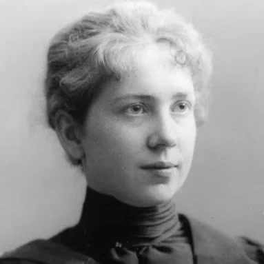 Black-and-white portrait of Harriet Brooks shown from the shoulders up. She faces slightly to the right with a neutral expression. Her hair is light-colored, loosely styled and swept back from her forehead. She wears a high-collared dark dress with a fitted neck and smooth fabric. The background is plain and light, with no visible objects or decorations.
