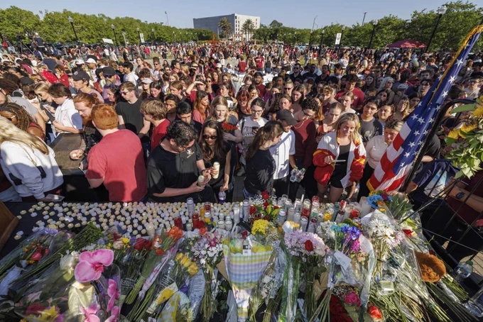 A large crowd of FSU students, faculty, and staff gathered on Langford Green for a solemn vigil remembering the tragedy of April 17, 2025. In the foreground, a memorial table is filled with dozens of lit candles and colorful bouquets of flowers, with attendees standing closely behind it.