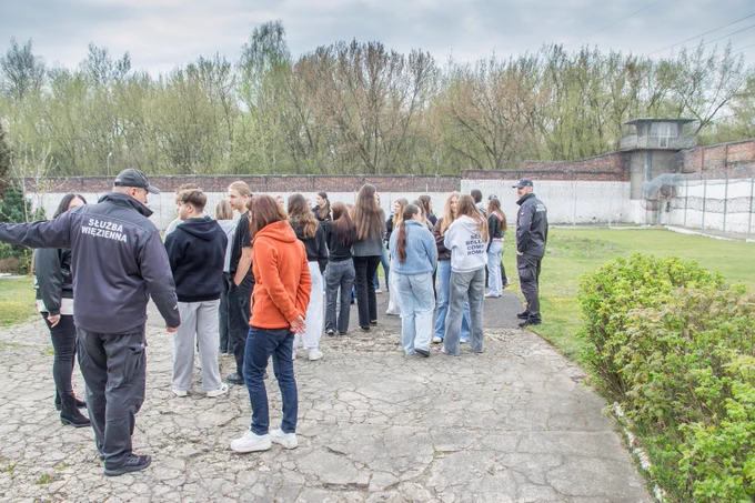 Grupa uczniów w cywilnych ubraniach ogląda teren ścisły jednostki penitencjarnej w asyście umundurowanych funkcjonariuszy Służby Więziennej.