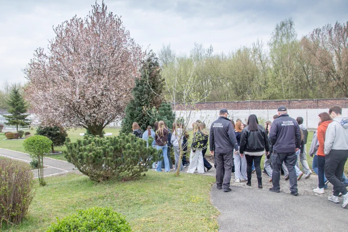 Grupa uczniów w cywilnych ubraniach ogląda teren ścisły jednostki penitencjarnej w asyście umundurowanych funkcjonariuszy Służby Więziennej.
