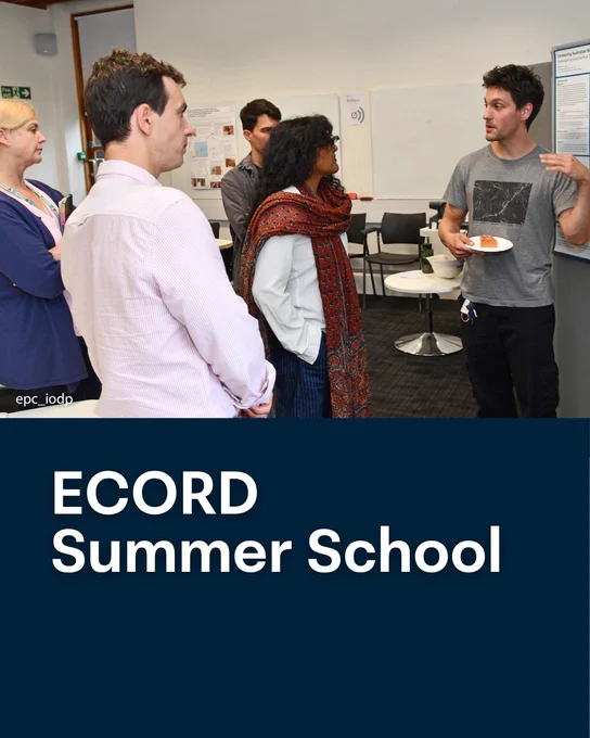 A group of people stand in conversation in a seminar room during the ECORD Summer School. One person holds a plate while speaking, others listen closely. Posters and chairs are visible in the background, indicating an academic networking or discussion setting.