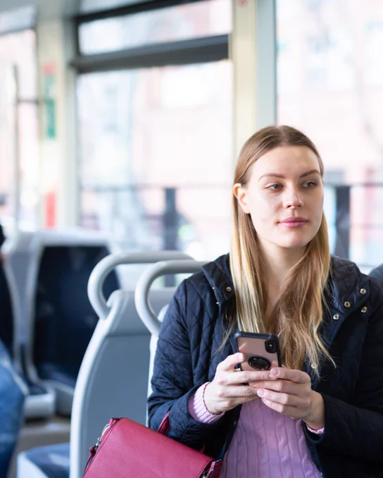 A woman on public transport holds her phone, looking away thoughtfully. She has blonde hair and wears a black jacket and pink jumper.
