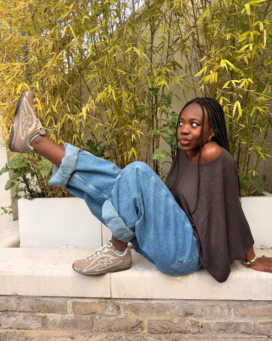 An image of a person wearing a pair of light brown New Balance 204L trainers with a summery outfit whilst sitting on a white stone wall in front of a yellow bush. 