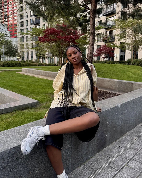 An image of a person wearing a pair of blue and grey New Balance 204L trainers whilst sitting on a concrete wall in a park.