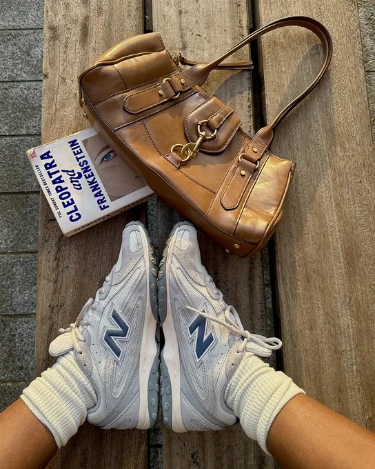 An image of a person's lower legs wearing a pair of grey and blue New Balance 204L trainers whilst resting their feet next to a book and a brown handbag.