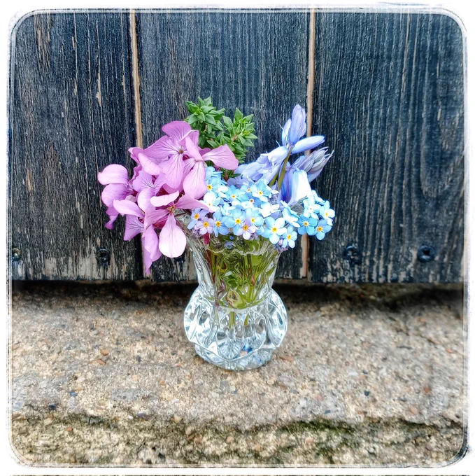 A little vintage glass vase filled with wild garden flowers, sat on a stone step with a dark painted wooden gate behind.