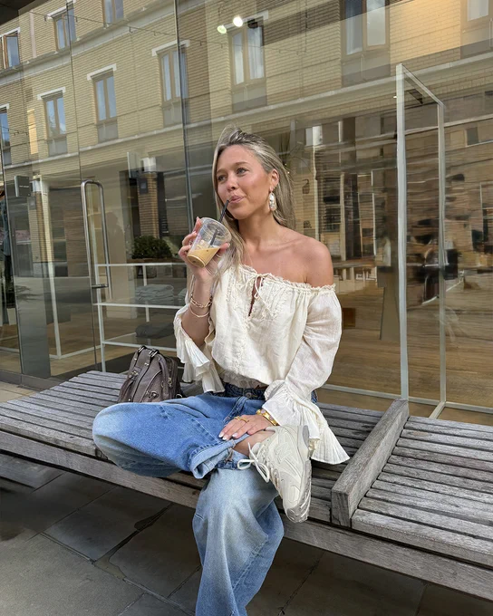 An image of a person wearing a pair of off white coloured New Balance 204L trainers with a summery outfit whilst sitting on a wooden bench and drinking a coffee.