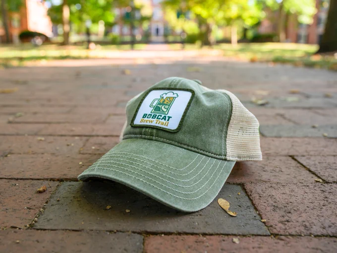 A green and beige baseball cap with the words "Bobcat Brew Trail" and a logo is lying on a brick pathway. The background features trees and a smooth pathway, softly blurred.
