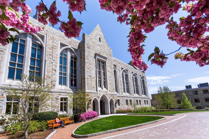 This image shows the exterior of McGuinn Hall at Boston College, framed by blooming pink cherry blossoms. A paved walkway leads to the entrance, and a person sits on a bench near vibrant flower bushes. The sky is clear and blue.