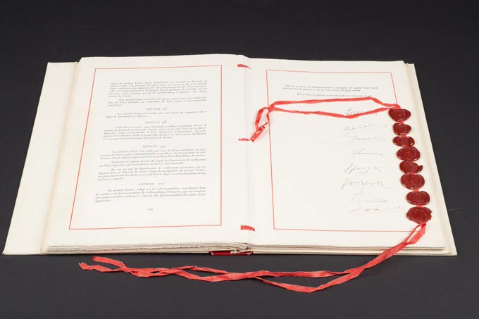 An open copy of the Paris Treaty lies on a dark surface, showing printed French text on the left page and a column of signatures on the right page, each accompanied by a red wax seal connected with red ribbons that trail out from the book. 