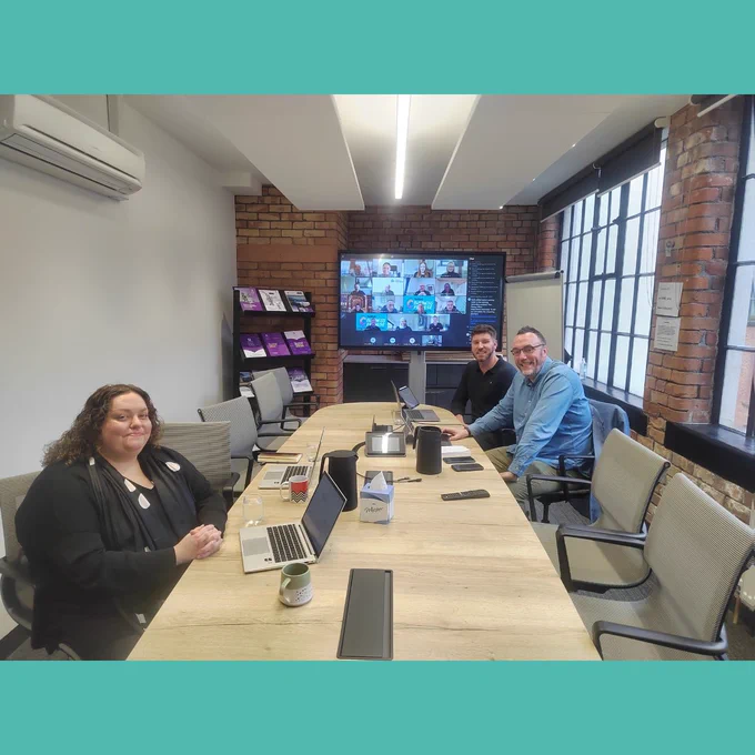 three people sit at a boardroom table smiling to camera, with a screen showing a video call with multiple participants behind them