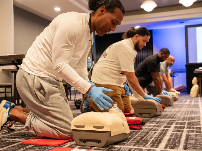 Participants kneel on a carpeted floor during a CPR training session, practicing chest compressions on mannequins. They wear gloves and are focused on the task. The setting appears to be a conference room with presentation equipment visible in the background.