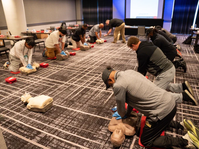 A group of people practicing CPR on mannequins in a classroom setting with a presentation screen at the front. Participants are kneeling on the floor, each working with a mannequin and a red training kit. The room is equipped with tables and chairs.
