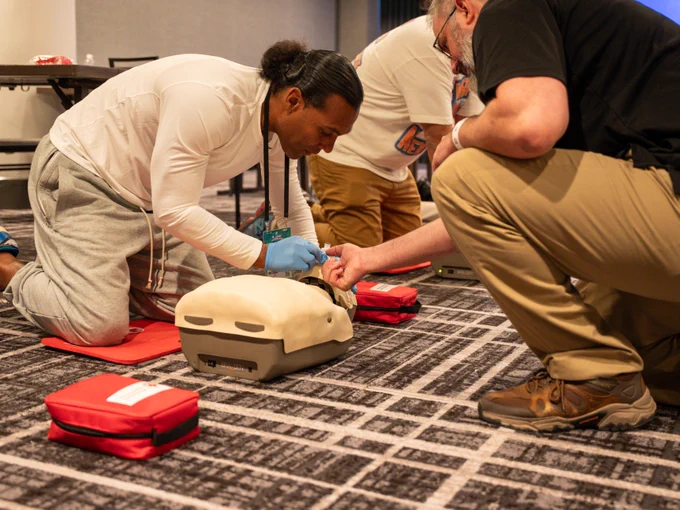 Two individuals are kneeling on the floor, practicing CPR on a training mannequin. One person is wearing blue gloves, and both are focused on the activity. Red first-aid kits are nearby on a patterned carpet.