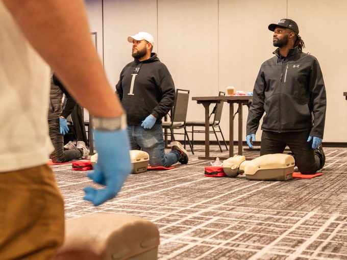 People are participating in a CPR training session. They are kneeling on the floor, each in front of a practice manikin. Participants are wearing gloves, and attention is focused on the training.