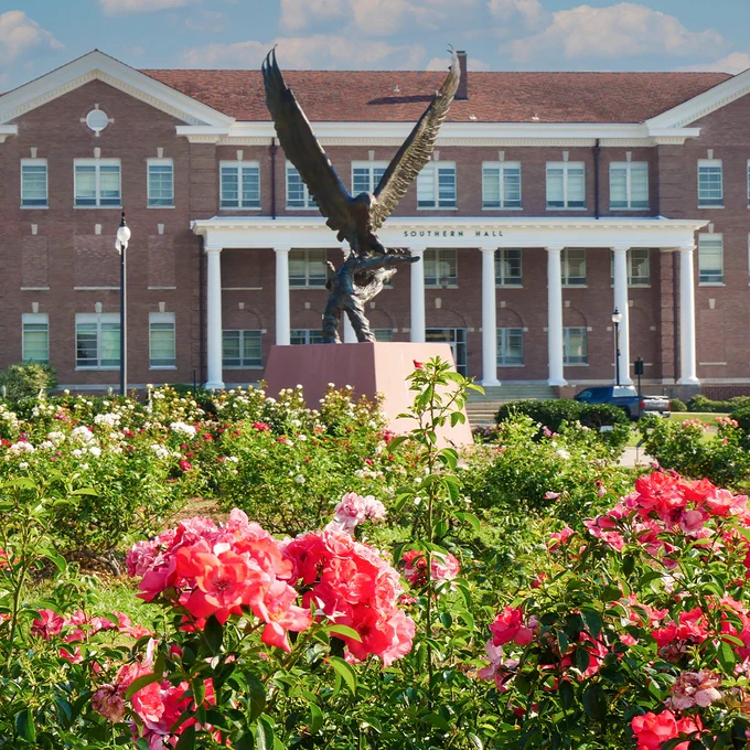 Pink and white flowers bloom in the foreground, with a large bronze eagle statue and a multi-story red brick building displaying the words "Southern Hall" in the background.
