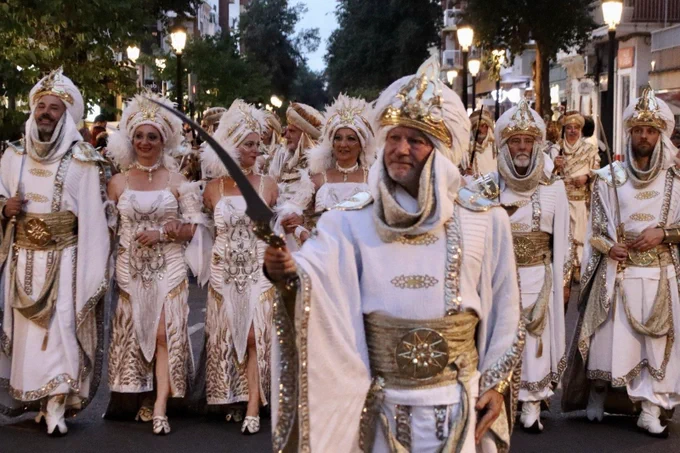 Desfile nocturno: personas con trajes blancos y dorados, turbantes y tocados; líder al frente blande un sable curvo.
