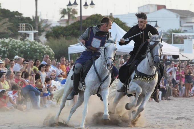 Dos jinetes vestidos de época chocan con lanzas cortas sobre caballos blancos en la playa mientras el público observa.