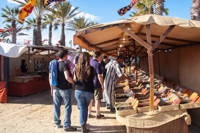 Mercado al aire libre junto a palmeras; puesto con cestas de especias y personas examinando productos bajo toldos.