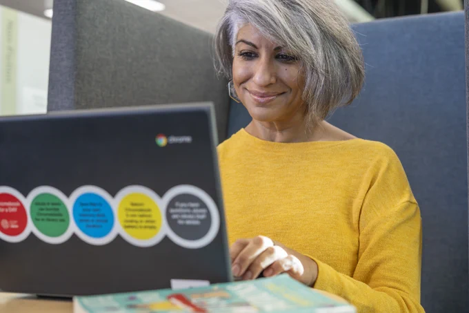 A person with gray hair is smiling while typing on a laptop with colorful circular stickers. The laptop has a Chrome logo. A book is partially visible on the table in front of the person.