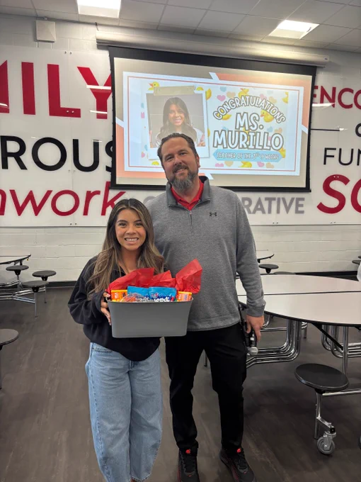 A smiling woman and a bearded man stand together holding a gift box in front of a congratulatory sign for "MS. MURILLO".
