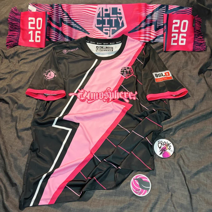 Minneapolis City SC kit which is black with a pink lightning bolt from the shoulder to the waist. Above it is a MPLS CITY SC scarf in shades of pink with white stripes and lightning bolts outlined in black and the years 2016 on the left and and 2026 on the right. There are two stickers bottom right with crows
