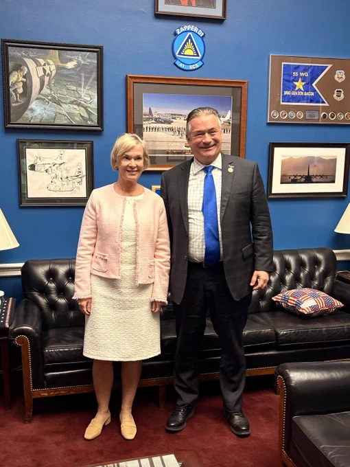 Ambassador and Senator standing in front of a dark couch and a blue wall filled with photos.