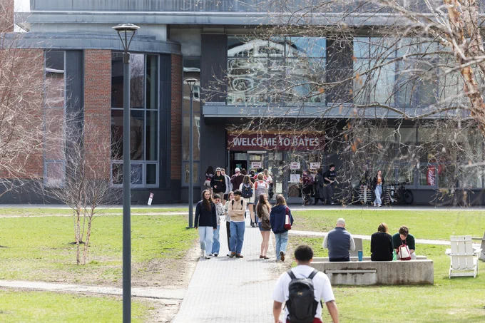 A group of prospective students walk away from Bates College's "Commons" dining hall on Alumni Walk. A "Welcome to Bates" sign can be seen in the background.