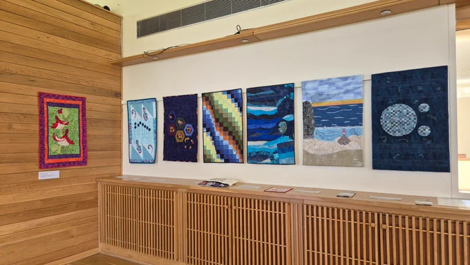Seven rectangular quilts hang side by side on a white wall, each with different patterns: abstract red and green shapes, light blue paisley motifs, multicoloured hexagons, a gradient of squares, an ocean scene, and circular planet like designs. A wooden cabinet sits below with small textile samples laid on top.