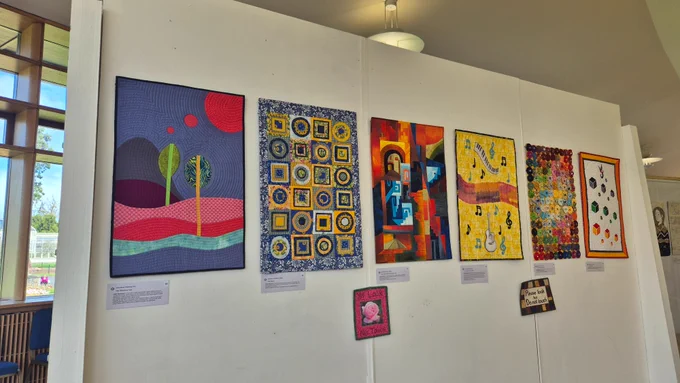 A row of six colourful textile artworks hangs on a white gallery wall. Each quilt has a distinct style, including abstract trees in purples and greens, blue and yellow geometric shapes, warm-toned angular architecture, musical motifs on yellow, rows of multicoloured circles, and a cube pattern design. 