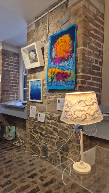 A stone and brick wall displays several textile and framed artworks. The largest piece is a bright abstract textile in orange, purple, blue, and yellow. Below it are two framed works: a soft-toned flower and a textured blue abstract. A lit white lamp with a felted shade sits to the right, illuminating small printed artist cards beneath it.