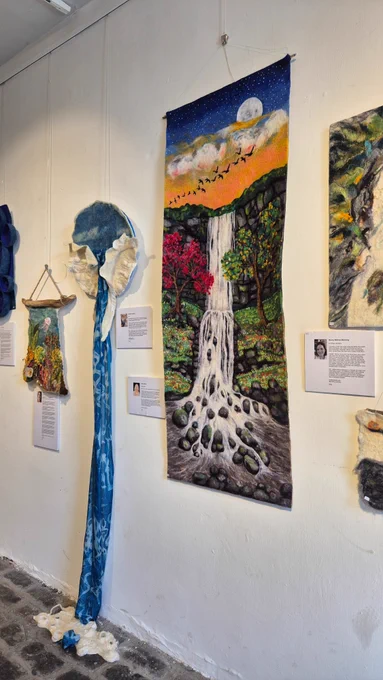 : A central textile artwork shows a waterfall flowing over rocks, surrounded by green foliage and red trees under a night sky with a full moon and birds. Above and below it hang additional textile pieces, including mountain and water themed designs. Each artwork has a small placard with text and an artist portrait.