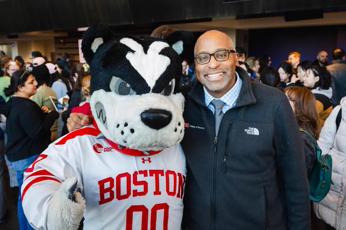 Boston University mascot Jack the Terrier, wearing a white hockey jersey with red \"BOSTON\" lettering and number 00, poses with College of Fine Arts Dean Harvey Young in a dark North Face zip jacket, light blue check shirt, and tie. The mascot gives a thumbs-up; both stand shoulder-to-shoulder in a busy indoor lobby with many blurred people and activity in the background.