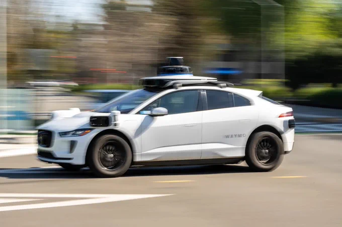A white Waymo autonomous vehicle, equipped with sensors on its roof and sides, is driving on a city street. The background appears blurred, indicating motion.