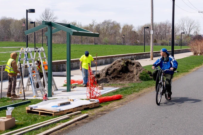 People installing a shade structure along a bike path while a person riding by on a bike watches.