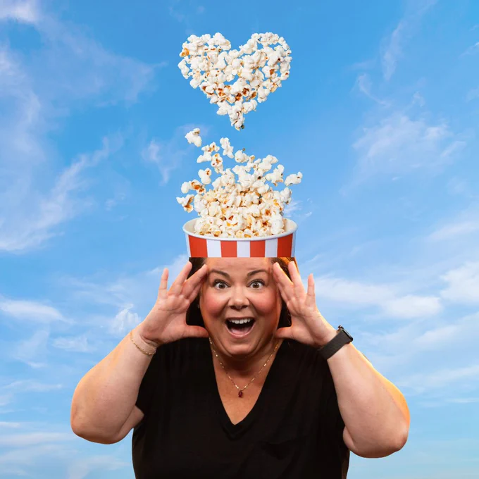 photo of me with both hands against my temples and a popcorn bucket "popping out" of the top of my head into a heart made out of popcorn in front of a blue sky.