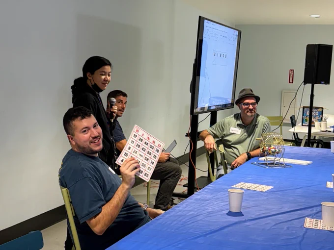 A group of people is gathered around a table covered with a blue cloth, participating in a bingo game. One person holds up a bingo card and appears to be smiling. A computer screen displays "Bingo" in the background. Cups are on the table and a bingo cage is visible next to the screen.
