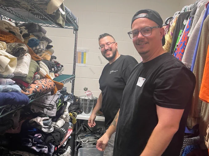 Two people are organizing clothes on shelves in a storage room. The shelves are filled with various folded clothes, and there are hangers with garments on the right side. One person is wearing glasses and casually dressed, and both appear to be engaged in their task.