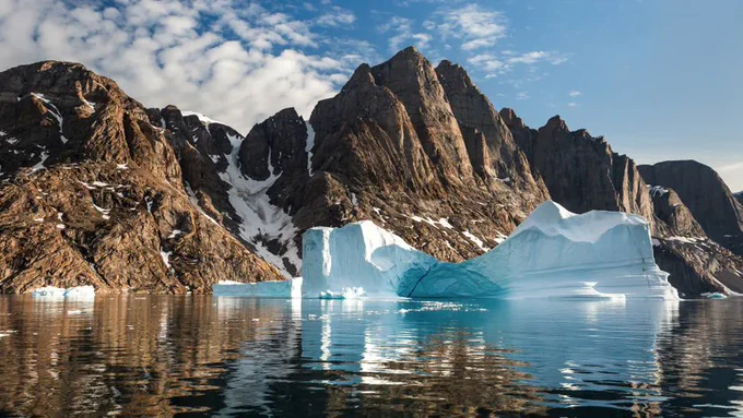 Clear blue water with a white glacier in the middle. Behind are large rocky mountains with some snow on them.