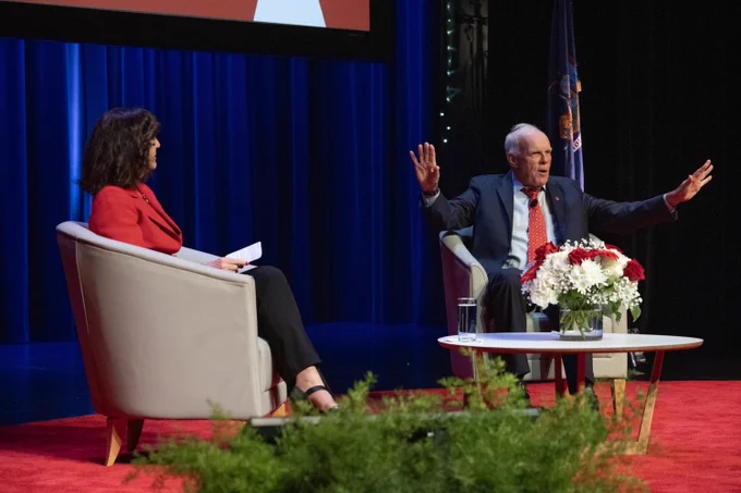 President Andrea Goldsmith and John L. Hennessy are seated on stage, engaged in conversation. A table with a floral arrangement is in front of them. A flag is visible in the background.