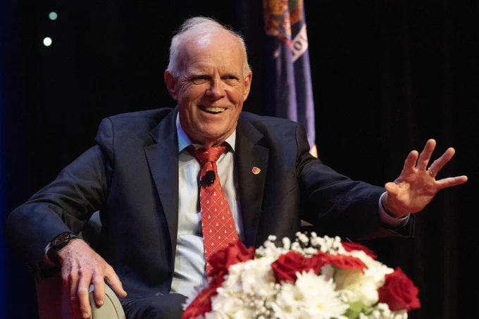 John Hennessy is sitting on stage during a conversation. He has his hand outstretched. 